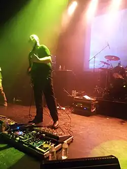 Der kahle Sänger steht auf einer in gelb getauchten Bühne, hält seine Gitarre in der Hand und trägt ein Headset in das er singt