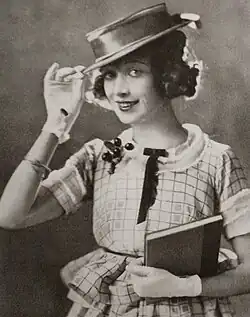 Esther Howard im Broadway's Musical The Sweetheart Shop 1920 lächelt und hat lockiges Haar. Sie trägt ein kariertes Kleid mit kurzen Ärmeln und einem Kragen. Am linken Kragen sind mehrere kleine, dunkle Kugeln angebracht, die wie eine Art Schmuck wirken. Sie hält in der linken Hand ein Buch und trägt weiße Handschuhe, die möglicherweise auf eine formelle oder traditionelle Situation hindeuten. Mit der rechten Hand hebt sie leicht einen breitkrempigen, karierten Hut an. Das Bild ist in Schwarz-Weiß gehalten.