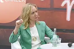 Foto einer Frau, die an einem runden Tisch sitzt und kritisch nach rechts blickt. Sie gestikuliert mit der rechten Hand.