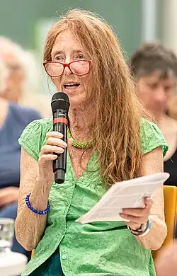 Eva Quistorp sitzt. Sie spricht in ein Mikrofon, das sie in der rechten Hand hält. In der linken Hand hält sie ein Schriftzug. Eva Quistorp hat schulterlanges rotes Haar, sie trägt eine rote Brille und eine grüne Bluse.