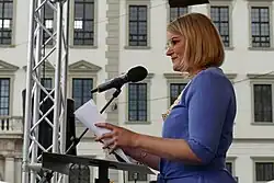 Oberkörper eine blonden Frau ein einem blauen Kleid, die an einem Stehpult mit Mikrofon steht. Hinter ihr ist die Fassade des Augsburger Rathauses zu erkennen.