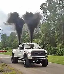 Das Bild zeigt einen Pick-up-Truck, der hinten zwei schwarze Rauchwolken ausstößt.