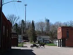 Das Ortszentrum von Fairchild, Wisconsin, mit einer weiten Straßenkreuzung, Einzelhäusern mit zwei Stockwerken aus Holz oder aus rötlichem Klinker und einem durchfahrenden Pferdefuhrwerk aus der Amish-Gemeinschaft