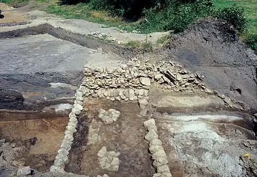 Ausgrabung 1983 (Blick nach Osten). Vorn die freigelegte Basis der Feldsteinmauer. Zwei Pfostengruben sind mit Feldsteinen gefüllt. Dahinter der Hauptwall im Profil, von links: Sodenwallreste, Feldsteinmauer, Mauerversturz über der Berme und Ansatz des einphasigen Grabens.