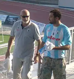 Maurizio Felugo ist auf der Tribüne eines Stadions zu sehen, im Hintergrund ist eine Laufbahn und eine Eckfahne erkennbar. Felugo trägt ein Hemd mit blau-weißen Längsstreifen und der Nummer 6 auf der Brust, dazu eine Tarnfarben-Hose. Pietro Figlioli ist während eines Spiels im Wasser, er trägt eine blaue Bademütze mit der weißen Nummer 4.