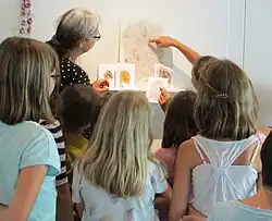 Nesa Gschwend mit einer Gruppe von Kindern in einem kreativen Umfeld. Sie stehen oder sitzen um Nesa Gschwend, die ihnen anscheinend Kunstwerke präsentiert. Sie hat graues Haar und trägt eine Brille. Sie deutet auf ein größeres Kunstwerk an der Wand, das aus Linien und Farben besteht – möglicherweise abstrakt. Vor diesem Kunstwerk befinden sich zwei kleinere Zeichnungen aufgestellt, die ebenfalls farbenfroh und linienbetont sind. Ein Kind hält eine Zeichnung in der Hand, die es den anderen zeigt. Die Szene strahlt eine pädagogische Atmosphäre aus und könnte in einer Kunstklasse oder einer Galerie aufgenommen worden sein, wo die Kinder sich mit kreativen Prozessen oder Kunstinterpretationen beschäftigen. Die Kleidung der Kinder ist lässig, und die Farben in der Umgebung sind harmonisch, was eine freundliche und offene Stimmung vermittelt.