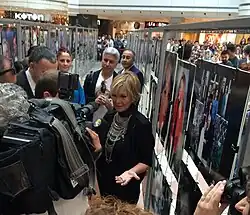 Filiz Akın mit blonden Haaren und auffälligem Schmuck spricht mit Journalisten vor einer Fotowand in einem Einkaufszentrum. Im Vordergrund steht Filiz Akın, die von Kameras und Mikrofonen umgeben ist und interviewt wird. Sie trägt schwarze Kleidung und eine auffällige Halskette. Um sie herum befinden sich weitere Personen, die an der Veranstaltung beteiligt zu sein scheinen. Der Hintergrund ist belebt: Viele Menschen bewegen sich durch das Einkaufszentrum, möglicherweise Besucher oder Teilnehmer dieser Veranstaltung. Auf beiden Seiten des Bildes stehen gerahmte Fotos oder Poster, die Teil einer Ausstellung sein könnten. Diese Bilder zeigen Porträts und Gruppenfotos. Außerdem ist der Name eines Geschäfts, „Koton“, sichtbar. Die Umgebung ist hell erleuchtet, und die Atmosphäre scheint lebendig und geschäftig, was auf ein besonderes Event hinweisen könnte.