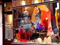 Frontale Farbfotografie von einem beleuchteten Schaufenster mit dunkler Holzumrandung. Zwei Schaufensterpuppen mit extravaganten Kleidern sind von Accessoires und einer glitzernden Leinwand umgeben. Auf dem Schaufenster ist die rote Silhouette von einem Teufel mit der goldenen Schrift „Wake up, Cave Girl“ abgebildet.
