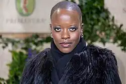 Florence Kasumba bei der Berlinale 2025. Sie trägt einen schwarzen Pelzmantel, der Eleganz und Stil ausstrahlt. Im Hintergrund sind grüne Pflanzen und ein Schild mit der Aufschrift „PRIMAVERA“ sowie ein Logo zu sehen, das wie ein grünes Wappen mit einer roten Figur darin wirkt. Sie steht im Mittelpunkt des Bildes und verkörpert Selbstbewusstsein.