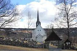 Foto einer weißen Holzkirche mit dunklem Kirchturm. Die Kirche ist von einem Friedhof umgeben, der wiederum von einer Steinmauer umgeben ist. Im Hintergrund eine hügelige Landschaft.
