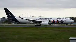 Seitenansicht eines Flugzeuges bei Start auf einer Landebahn. Auf dem Flugzeug sind abstrakt überwiegend Menschen verschiedener Hautfarben mit Deutschlandtrikots. „Diversity Wins. Lufthansa“ steht darüber.
