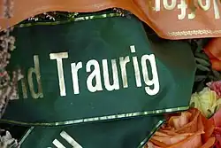 Das Wort „Traurig“ in goldener Schrift auf einem dunkelgrünen Stoff, am Rande des Bilds sind weitere Trauerschleifen in anderer Farbe und Zweige und Blumen zu sehen.