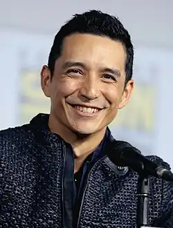 Gabriel Luna auf der Comic-Con in San Diego. Er trägt eine dunkelblaue, strukturierte Jacke mit einem schwarzen Reißverschluss und Knöpfen. Vor ihm befindet sich ein Mikrofon, was darauf hindeutet, dass er möglicherweise eine Rede hält oder an einer Diskussion teilnimmt. Im Hintergrund ist ein unscharfes Banner zu erkennen, das vermutlich das Logo der Comic Con zeigt.
