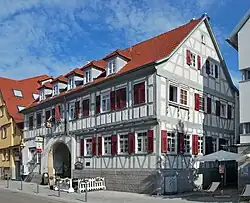 Ein zweistöckiges, unverputztes Fachwerkgebäude mit weißem Gefache und grau gestrichenen Balken und mit einem roten Ziegeldach mit Gauben