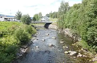 Foto eines Flusses, in dem sich mehrere Felsen befinden. Über den Fluss führt eine Brücke und entlang des Flusses befinden sich einige Gebäude.
