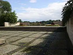 Der vordere Bereich ist durch Stufen vom hinteren etwas abgesetzt. Dort hinten gibt es zwei quadratische Blumenbeete und ein rotes Dreieck mit Spitze nach unten an einem Geländer, dahinter der große Teich