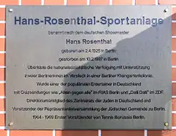 Gedenktafel, die Hans Rosenthal gewidmet ist. Mit der Aufschrift Hans-Rosenthal-Sportanlage. Die Tafel ist aus Metall und wurde auf rotem Ziegelstein angebracht, was einen Kontrast zum Hintergrund bildet. Die Inschrift enthält einen Überblick über Rosenthals Leben und Verdienste, wobei seine Geburts- und Sterbedaten angegeben sind. Es wird auch darauf hingewiesen, wie er die nationalsozialistische Verfolgung überlebte, indem er sich versteckte, und später zu einem bekannten Entertainer in Deutschland wurde. Die Tafel würdigt außerdem seine Rolle in jüdischen Organisationen und seinen Beitrag zur Gesellschaft. Die Tafel vermittelt nicht nur eine Erinnerung an seine Leistungen, sondern auch seine Bedeutung als Persönlichkeit, die schwierige Zeiten überwunden hat.