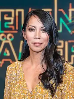 Minh-Khai Phan-Thi bei der Aufzeichnung des Westdeutschen Rundfunks im Studio Köln-Bocklemünd vom 24. Januar 2018 mit langen dunklen Haaren, die ein gelbes Kleid mit Blumenmuster trägt. Der Hintergrund ist bunt und abstrakt, mit Text und Formen von der Sendung "Geheimniskrämer" mit Martin Klempnow und seinen Gästen.