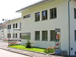 Gebäude mit heller Fassade, davor auf einer Glasstele das Wappen der Gemeinde, darüber die Aufschrift „Gemeindeamt St. Gotthard“, darunter „Hort“ (für die Kinderbetreuungsstätte im Untergeschoß des Gebäudes)