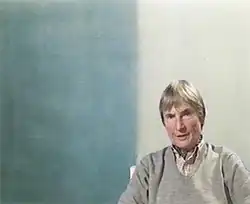 Eine ältere Frau mit kurzen grauen Haaren und schmalem Gesicht sitzt vor einem Ölgemälde, das vertikal in zwei Farbflächen geteilt ist. Links etwas breiter ein helles blau, rechts ein neutralerer, fast weißer Ton. Die Frau sitzt rechts vor dem hellen Farbton, nur leicht mit der Schulter in den blauen hineinragend