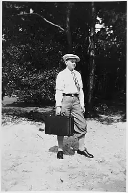 Die Schwarz-Weiß-Fotografie zeigt eine bartlose junge Person in Hemd und Hosen mit einer Mütze auf dem Kopf und einem Aktenkoffer in der rechten Hand.