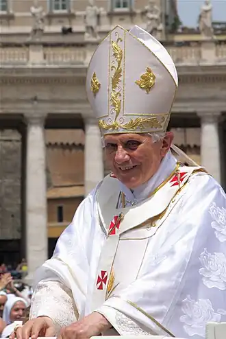 Farbfotografie Benedikts XVI. in liturgischer Kleidung und Mitra aus Stoff mit goldenen und roten Ornamenten. Im Hintergrund sind ein Gebäude und Menschen zu sehen.