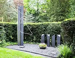 Eine Grabstätte. auf dem kiesbedeckten Untergrund stehen vier etwa kniehohe Dreiecksäulen aus schwarzem Stein, deren Deckflächen in verschiedenen Richtungen abgeschräng sind. Links daneben eine etwa 2,50 m moderne Plastik aus zwei sehr schmalen Quadern und diese chaotisch durchbrechenden dicken Metalldrähten. Beschriftung auf zwei Prismen: Mona Scholl 1966 1981, Rudolf Scholl, 1931 2018