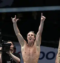 Grant Brits jubelt mit nacktem Oberkörper vor dunklem Hintergrund, am rechten Bildrand ist ein Kameramann zu sehen.