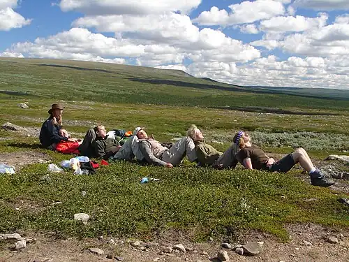 6 Personen sitzen hintereinander auf einer weitläufigen Tundra (jeweils an die aufgestellten Schienbeine der hinteren Person gelehnt) und genießen die Sonne