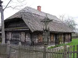 Farbige Eckansicht eines dunklen Holzhauses mit Reetdach und einem roten Schornstein. Die Fensterläden sind geschlossen. Im Vordergrund sind ein grauer Lattenzaun und ein Jesuskreuz mit einer betenden Figur aus Holz darunter.