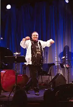 Gustav Peter Wöhler 2005 auf einer Bühne, seines Konzertes mit seiner Band. Er trägt ein weißes Hemd über einem schwarzen T-Shirt mit einem teilweise sichtbaren Schriftzug („sexy“). Er hat beide Arme erhoben, was auf Bewegung oder Interaktion hinweist. Im Hintergrund ist ein blauer Vorhang zu sehen, der die Bühne umrahmt.Die Bühne selbst ist mit verschiedenen Musikinstrumenten ausgestattet. Dazu gehören ein großes Cello oder Kontrabass, ein Klavier und mehrere Notenständer mit Notenblättern. Es sind zudem Mikrofone und Lautsprecher vorhanden, die auf eine musikalische Aufführung hinweisen. Die Beleuchtung lenkt die Aufmerksamkeit auf die Person und die Bühne.