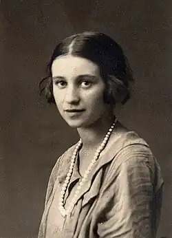 Junge Frau mit sanftem Blick und leisem Lächeln, halblange Haare mit Mittelscheitel, in einem gefälteten Kleid mit einer Perlenkette