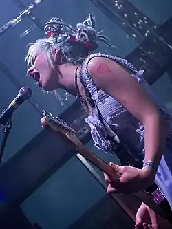 Auf dem Bild ist Lauren Tate im Profil zu sehen. Sie wurde hier bei einem Auftritt ihrer Band Hands of Gretel fotografiert und steht mit einer E-Gitarre am Mikrofon und singt oder schreit dort hinein.