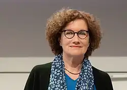 Porträt einer Frau mit gewelltem, etwa kinn- bis nackenlangem Haar, Brille mit oval-runden Gläsern und einem blau-gemusterten Schal um den Hals