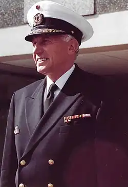 Ein älteres Bild eines Marineoffiziers. Er trägt eine Kapitänsmütze, die sein graues Haar verdeckt und guckt leicht lächelnd nach links vorne. Oberhalb der linken Brusttasche seines Zweireihigen Sakkos befinden sich einige Auszeichnungen. Unter dem Sakko trägt er ein weißes Hemd und eine schwarze Krawatte.