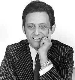 Autogrammkartenmotiv zeigt Hans Rosental, der einen Nadelstreifenanzug und eine Krawatte trägt. Er lächelt und stützt eine Hand nachdenklich auf das Kinn, wobei die Armbanduhr am Handgelenk sichtbar ist. Das Bild ist in Schwarz-Weiß gehalten und vermittelt ein elegantes, formales Ambiente.