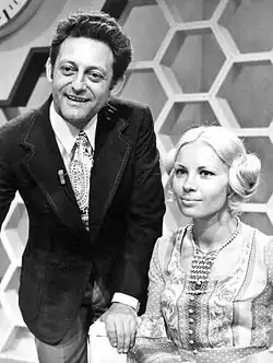 Hans Rosenthal und Monika Sundermann in der Sendung Dalli-Dalli (1971) Hans Rosenthal trägt einen dunklen Anzug mit einer gemusterten Krawatte.Er hat dunkles, welliges Haar und steht leicht nach vorne gebeugt. Monika Sundermann auf der rechten Seite sitzt und trägt ein Kleid mit einem komplexen Muster sowie eine Perlenkette. Ihr Haar ist hell und zu einem stilvollen Knoten hochgesteckt. Der Hintergrund besteht aus einer Wand mit sechseckigen Muster. Diese Muster waren typisch für die Sendung Dalli Dalli.