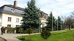 Das Bild zeigt ein weißes Gebäude mit schwarzem Dach, das etwa zur Hälfte von Bäumen verdeckt ist.