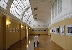 ein Gang mit Sprossenfensteroberlicht links. Die Wände sind bis etwa mannshoch ocker-orange, darüber weiß. Soweit im oberen Bereich keine Fenster sind, zieht sich dort ein Streifen an Schablonenmalerei entlang