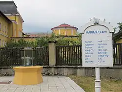 Links ein Glaskasten mit stark sprudelndem Wasser auf einem gelben Sockel, rechts ein Schild mit der Aufschrift Marian D IVX und den Werten der Quelle