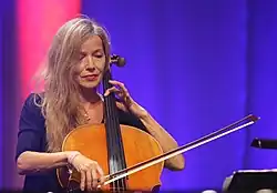 Fany Kammerlander (2016), die ein Cello spielt. Sie hat lange, blonde Haare, die offen fallen, und trägt ein dunkelblaues Oberteil. Sie hält den Bogen mit der rechten Hand und greift mit der linken Hand die Saiten des Cellos, das vor ihr steht. Der Hintergrund des Bildes ist in einem sanften Lila beleuchtet, was darauf hinweist, dass die Aufnahme möglicherweise bei einer Bühnenaufführung oder einer Probe gemacht wurde. Zudem trägt die Person Schmuck, darunter ein Armband am linken Handgelenk und eine zarte Halskette mit einem kleinen Anhänger.