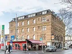 Straßenansicht eines mehrstöckigen Backsteingebäudes an einer belebten Kreuzung in einem urbanen Umfeld. Im Erdgeschoss befinden sich Geschäfte, darunter eine Apotheke. Vor dem Gebäude stehen geparkte Autos und mehrere Personen gehen auf dem Gehweg. Links im Bild ist ein Imbissstand mit rotem Dach und Menschen davor zu sehen. Ein Straßenschild und ein Laternenmast sind ebenfalls sichtbar. Im Hintergrund sind weitere Gebäude und eine Bahnunterführung erkennbar. Der Himmel ist leicht bewölkt.