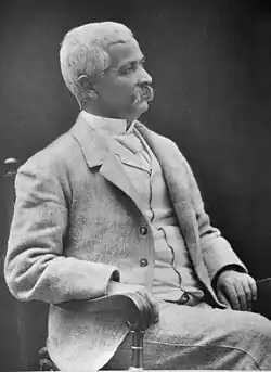 Eine Alte Aufnahme ist zu sehen von Henry Morton Stanley, mit seinem typischen Schnurrbart. Dieser sitzt in einem Stuhl, sichtlich gealtert mit bereits weißen Haar und trägt einem hellen Anzug.