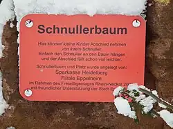 Text auf der Tafel: „Schnullerbaum. Hier können kleine Kinder Abschied nehmen von ihrem Schnuller. Einfach den Schnuller an den Baum hängen und der Abschied fällt schon viel leichter. Schnullerbaum und Platz wurde angelegt von: Sparkasse Heidelberg, Filiale Eppelheim, im Rahmen des Freiwilligentages Rhein-Neckar 200x (letzte Ziffer teilweise von Schnee bedeckt) mit freundlicher Unterstützung der Stadt Ep… (Rest von Schnee bedeckt)“