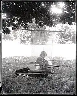 Schwarz-weiß-Foto von We’whas Arbeit an einem Webstuhl. We’wha sitzt dabei auf einer Wiese. We’wha trägt ein bis zu den Füßen reichendes Gewand, dessen Ärmel weiß sind, der Rest ist hingegen schwarz. We’wha trägt eine ähnliche Halskette wie vorher, diesmal stecken in einigen der Steinchen Stacheln. Hinter beziehungsweise über We’wha sind Baumblätter und Bäume zu sehen.