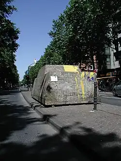 Ein grauer Betonklotz mit erkennbarer Form eines Autos auf einer Verkehrsinsel einer mehrspurigen Straße, Erkennbar sind Graffitis, gelbe Farbe