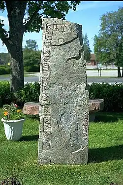 Im schrägen Sonnenlicht ist die Runeninschrift gut erkennbar, die als Band entlang des Randes der Vorderseite eines annähernd quaderförmigen, aufrecht stehenden Steins eingeritzt wurde, aber durch eine großflächige Beschädigung der Steinoberfläche unterbrochen wird; im Hintergrund ein Blumenkübel und eine Steinbank unter einem Baum