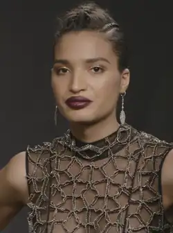 Porträtfoto von Indya Moore. Moore trägt dunkelroten Lippenstift, goldenen Lidschatten, ein dunkelbraunes Top mit einem diamantbesetzten Netzmuster und an beiden Ohren silberne Ohrringe mit an Ketten hängenden Federn. Die braunen Haare trägt Moore als bis zu den Ohren reichende, nach oben abstehende Kurzhaarfrisur.