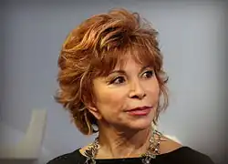 Isabel Allende auf der Frankfurter Buchmesse 2015, mit kurzen, welligen Haaren, die in verschiedenen Rottönen gefärbt sind, von dunkleren bis hin zu helleren Nuancen. Sie trägt eine auffällige Halskette mit großen silbernen und transparenten Anhängern, die einen interessanten Kontrast zum schlichten grauen Hintergrund bildet. Im Hintergrund ist zudem ein Teil eines weißen Stuhls sichtbar, der jedoch nur am Rand des Bildes erscheint und keine weiteren auffälligen Details zeigt. Die Gesamtkomposition des Bildes wirkt ruhig und fokussiert auf die Person.