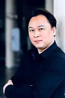 James Lee (2019) trägt ein schwarzes Hemd und hat die Arme vor der Brust verschränkt. Er steht vor einer hellen Wand, und der Hintergrund ist leicht verschwommen. Es scheint sich um eine Innenaufnahme zu handeln, wobei im Hintergrund einige vertikale Strukturen und möglicherweise ein Schild oder Poster sichtbar sind. Er hat dunkles Haar und ist leicht rechts von der Bildmitte positioniert. Die Pose der Person wirkt entspannt und selbstbewusst.